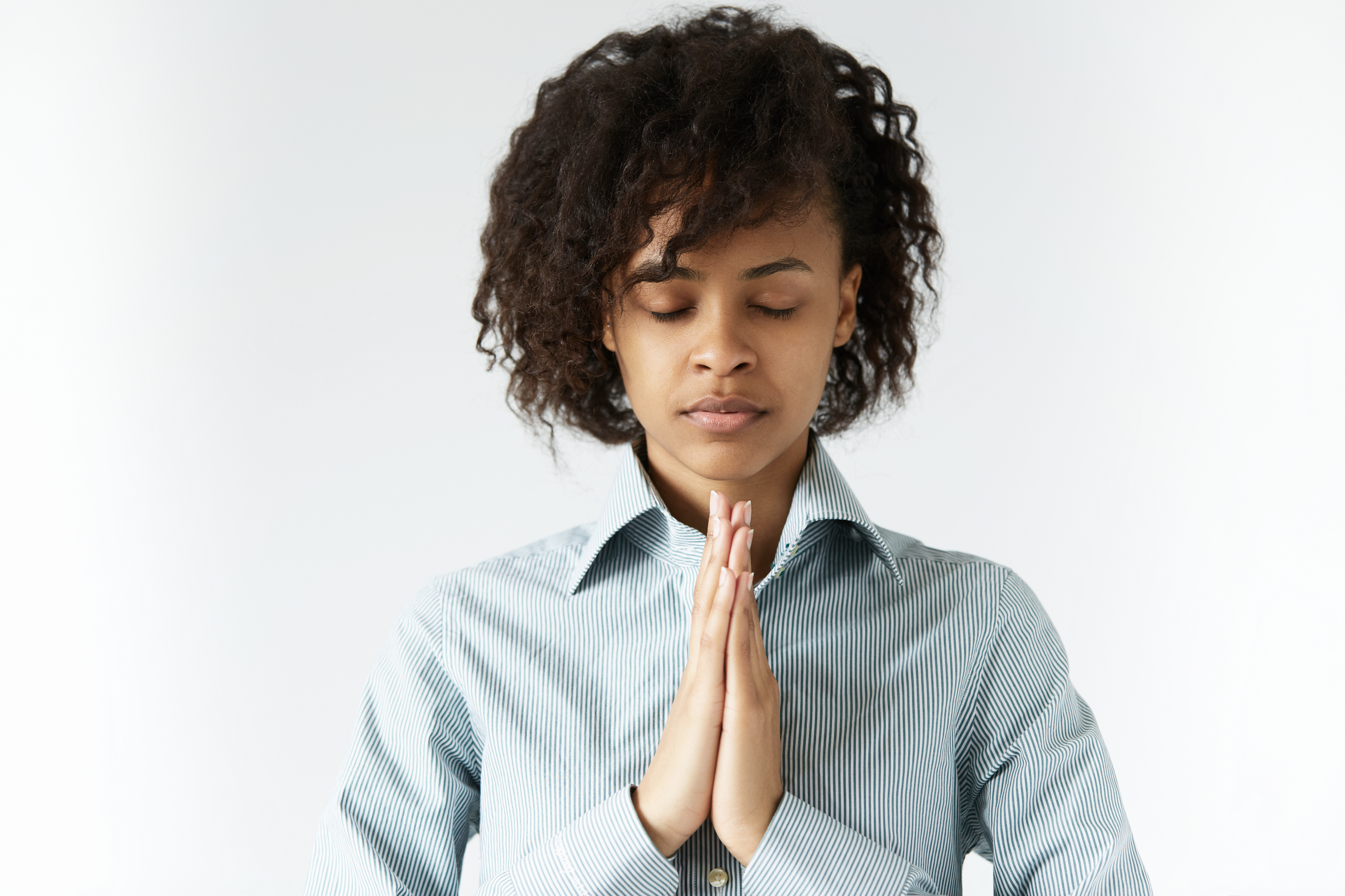 Concept of meditation and prayer. African female standing in meditative pose against white wall, with closed eyes and calm face full of sense of inner peace and wisdom, holding hands in prayer